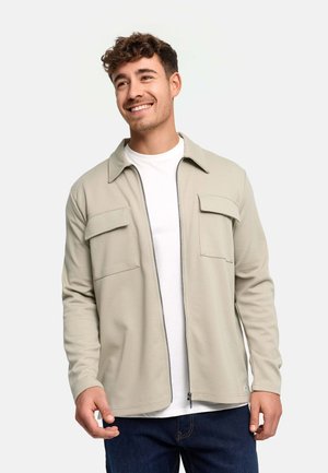 Homme souriant portant une veste beige zippée sur une chemise blanche et un jean foncé, debout devant un fond clair uni.
