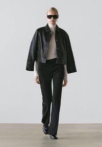 Woman walking in black leather jacket over beige turtleneck, black pants, black shoes, and large black sunglasses against plain background.