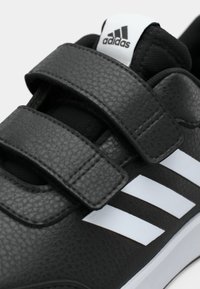 Crne Adidas tenisice od teksturirane kože, s dva čičak traka, bijelim Adidas logotipom na jeziku i tri bijele bočne pruge.