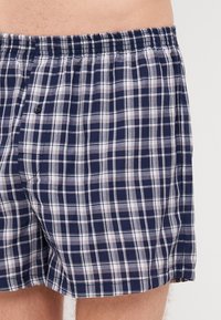 Boxers azul-marinho e brancos com padrão quadriculado, com um cós elástico e um único botão. Material de algodão, corte relaxado. Tecido levemente texturizado.