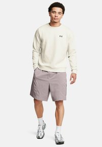 Hellbeige Sweatshirt mit gerippten Bündchen, kombiniert mit helllila Shorts mit seitlichen Taschen. Die Schuhe sind grau mit weißen Akzenten und Schnürsenkeln.