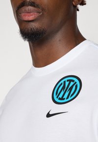 Hvid T-shirt med kortærmer, der har et sort Nike-logo og et blåt Inter Milan-emblem. Blødt stof med rund halsudskæring og uden synlige mønstre.