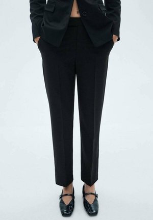 Personne portant un pantalon noir cintré, un blazer noir les mains dans les poches, et des chaussures noires à talons bas sur un fond blanc uni.