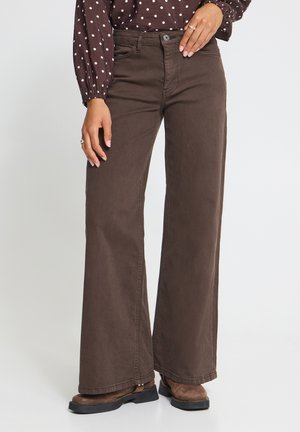 Vrouw die bruine wijde broekspijpen draagt, bruine laarzen en een bruine blouse met lange mouwen en witte stippen, staand met één hand op haar heup.