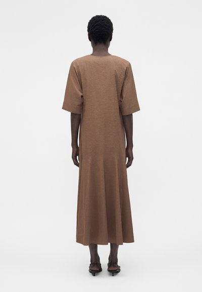 Άτομο με κοντά σγουρά μαλλιά φορά χαλαρό καφέ μίντι φόρεμα και μαύρα σανδάλια με χαμηλό τακούνι, στέκεται με την πλάτη στραμμένη προς τα εμπρός.