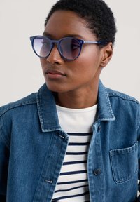 Blå kattögon-solglasögon med gradientlinser, bärs tillsammans med en jeansjacka och en randig T-shirt. Solglasögonen har tunna bågar och rundade kanter.