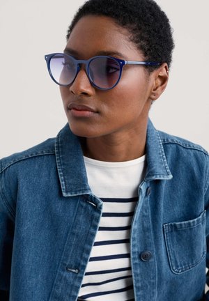Blue cat-eye sunglasses with gradient lenses, worn with a denim jacket and a striped T-shirt. The sunglasses feature thin frames and rounded edges.