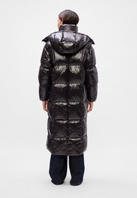 Geacă puffer neagră cu un exterior lucios, glugă supradimensionată și un design până la genunchi. Prezintă secțiuni matlasate și o siluetă asimetrică la talie.