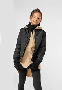 Svart quiltet bomberjakke over en beige zip-up hettegenser, kombinert med svarte votter og leggings. Glatt stoff, ribbede mansjetter og høy krage.