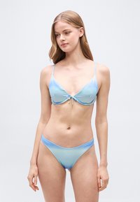 Νεαρή γυναίκα που φοράει μπικίνι σε ανοιχτό μπλε ombre με λεπτούς ιμάντες, στέκεται μπροστά σε απλό φωτεινό γκρι φόντο.