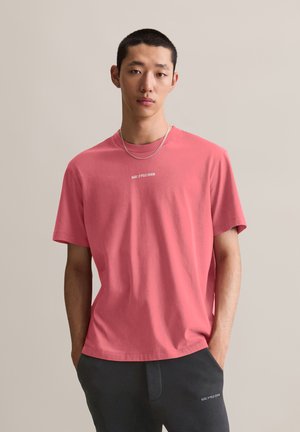 Kurzärmliges, Rundhals-T-Shirt aus korallfarbenem Baumwollstoff mit einem dezenten weißen Logo-Druck auf der Vorderseite. Sanfte Textur und lässige Passform.