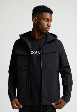 Hombre con cabello rizado corto, llevando una chaqueta negra sobre una camiseta negra, mirando a su derecha, sosteniendo los lados de la chaqueta cerca de la cremallera.