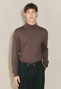 Brauner Rollkragenpullover mit langen Ärmeln, weicher Textur und geripptem Kragen, kombiniert mit schwarzen Hosen und einem schwarzen Gürtel.