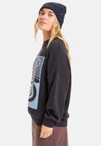 Svarte oversized genser med et blått grafisk mønster, med ribbestrikkede ermer og kant. Brukt med en matchende marineblå lue. Glatt tekstur.
