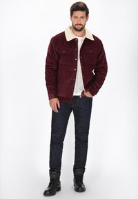 Bordeaux fløjlskursted med en beige faux shearling krave, to brystlommer, knaplukning og afslappet pasform. Båret over en hvid skjorte og blå jeans.