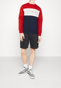 Sudadera roja y marina con logo, pantalones cortos de cargo negros y zapatillas grises con acentos blancos. Material de mezcla de algodón, corte relajado.