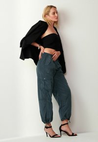 Čierny crop top, voľné teplákové nohavice v odtieni teal s bočnými vreckami a čierne pásikové lodičky. Model pózuje na bielom pozadí.