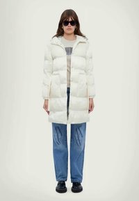 Λευκό παλτό με επένδυση και κουκούλα, διαθέτοντας κλείσιμο με κουμπιά και πλαϊνές τσέπες, φορεμένο πάνω από ένα γκρι μπλουζάκι και μπλε τζιν με σχέδιο λογότυπου.