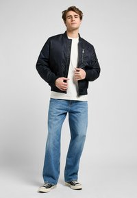 Jeune homme portant une veste bomber noire, un t-shirt blanc, un jean bleu et des baskets noires, se tenant devant un fond gris uni.