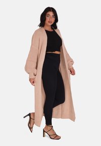 Lång, stickad cardigan i mjuk beige med en avslappnad passform, som har överdimensionerade ärmar och en subtil textur, i kombination med svarta leggings och sandaler med remmar.