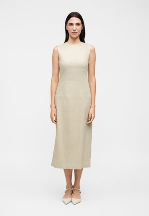 Γυναίκα με μακριά σκούρα μαλλιά που φοράει αμάνικο μπεζ φόρεμα μήκους μίντι και ανοιχτόχρωμα μυτερά παπούτσια, στέκεται μπροστά σε απλό λευκό φόντο.