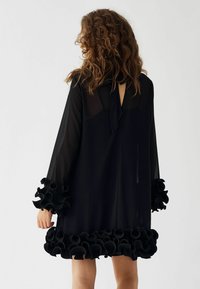 Črna prosojna obleka z dolgimi rokavi, ki ima naborke na manšetah in robu, z vezanim izrezom na vratu ter ohlapnim krojem.