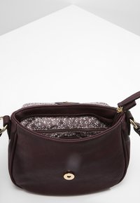 Hnědá kožená kabelka přes rameno se zlatým zapínáním, květinovou látkovou podšívkou a zipovými přihrádkami, položená na bílé ploše.