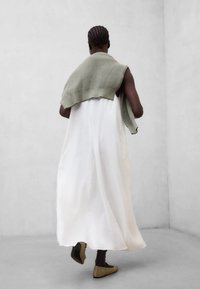 Personne aux cheveux tressés portant une longue robe blanche, un pull vert olive posé sur les épaules, et des chaussures beige, marchant dans un espace gris minimaliste.
