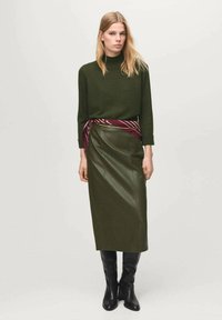 Maglione verde oliva con collo alto, abbinato a una gonna di pelle verde scuro con spacco laterale, accentuata da una cintura a motivi bordeaux. Stivali neri sopra il ginocchio.