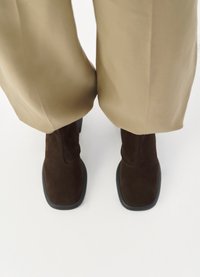 Brown suede ankle boots with a matte finish, featuring a black rubber outsole and minimal stitching, positioned beneath beige wide trousers.