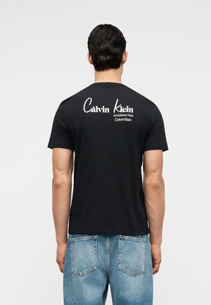 Homme portant un t-shirt noir Calvin Klein avec le texte de logo blanc "Calvin Klein Established 1968" dans le haut du dos, associé à un jean bleu clair.