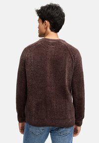 Man met kort donker haar, gekleed in een gestructureerde donkerbruine trui en blauwe jeans, met zijn rug naar de kijker gekeerd tegen een effen lichtgrijze achtergrond.