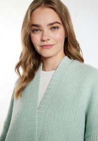 Světle máta zelený kardigan s texturovaným pleteným designem, s hlubokým výstřihem do V a žebrovanými detaily po obvodu a na okrajích.