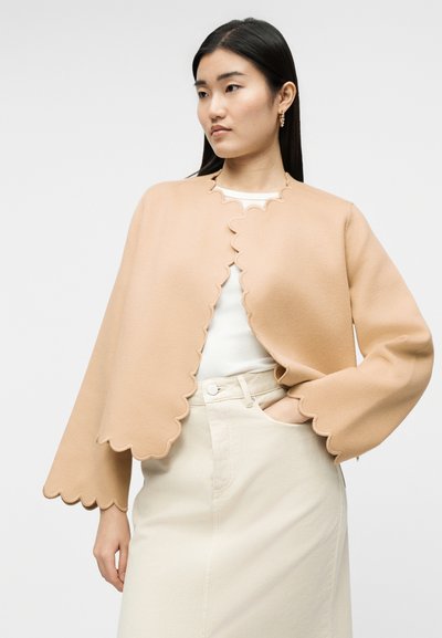 Γυναίκα που φορά ένα μπεζ μπουφάν κάπα με κυματιστό τελείωμα πάνω από λευκή μπλούζα και μπεζ φούστα ψηλόμεση, στέκεται έχοντας το ένα χέρι στην τσέπη.