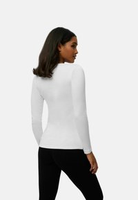 Top blanco de manga larga hecho de una tela suave con un diseño ajustado. Presenta un cuello redondo y un dobladillo curvado. Combinado con leggings negros.