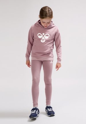 Ung pige i lilla hoodie og leggings med et hvidt logo, kigger ned, iført marineblå og hvide sneakers, stående mod en ensfarvet baggrund.