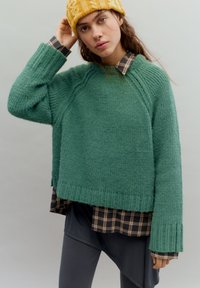 Vihreä neulottu neule, jossa on resorireunus helmassa ja hihoissa, kerrospukeutuneena tummanruutuisen paidan päälle. Mallilla on keltainen neulottu myssy. Pehmeä, teksturoitu kangas.