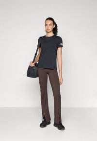 Svart T-skjorte med korte ermer, brune split-bukser og svarte joggesko. Modellen holder en liten svart tilbehørspose. Enkelt design og passform.