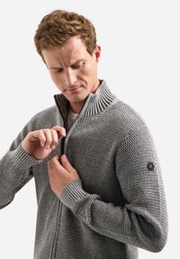 Grijze gebreide ritstrui met een gestructureerd patroon, geribbelde kraag en manchetten, en een logopatch op de linkermouw. Model past de rits aan.