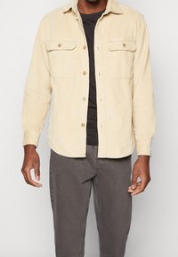 Beżowa sztruksowa koszula-żakiet z dwoma kieszeniami na piersiach, zapinana na guziki, z długimi rękawami, noszona na czarnej koszulce, o fakturowanej tkaninie.