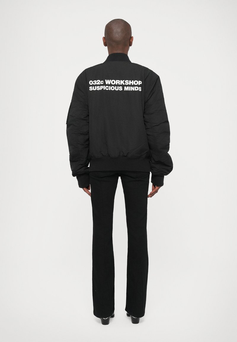 Person standing facing away wearing black jacket with "032c WORKSHOP SUSPICIOUS MINDS" text on back, black pants, and heeled shoes.