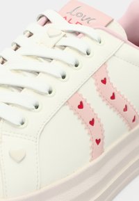 Baskets blanches avec accents roses, découpes en forme de cœur sur une superposition rose, semelle en caoutchouc texturée et porte-lacets en forme de cœur.