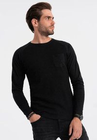 Uomo in camicia nera a maniche lunghe con taschino e jeans neri, che indossa un orologio e braccialetti, che guarda di lato su uno sfondo semplice.