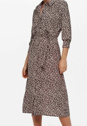Frau trägt ein braunes Midikleid mit kleinen weißen Blumenmustern, dreiviertel Ärmeln, einem Kragen und einer taillierten Passform.