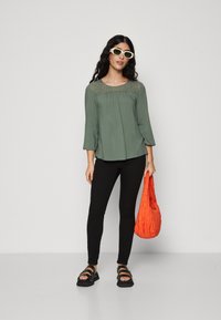 Grønn bluse med blonderdetaljer på skuldrene, trekvart ermer, sammen med svarte leggings og kraftige svarte sandaler. Oransjepose.