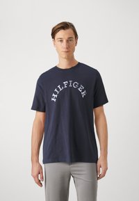 Marineblå bomull T-skjorte med korte ermer, med teksten "HILFIGER" i en buet hvit design over brystet. Crew-hals.