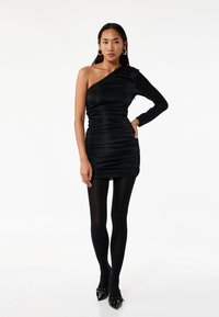 Robe mini noire à une épaule avec texture froncée, présentant une manche longue d'un côté et une coupe ajustée. Portée avec des collants noirs et des chaussures pointues.