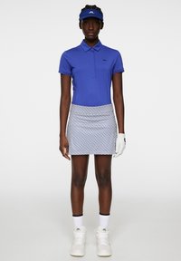 Blå polo skjorta med korta ärmar, vit mönstrad kjol, vita träningsskor och en matchande basker. Golfhandske på vänster hand.