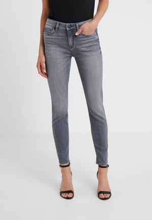 Femme portant un jean skinny gris et des talons noirs à bout ouvert avec sangle à la cheville, debout avec une main dans la poche devant un fond uni.