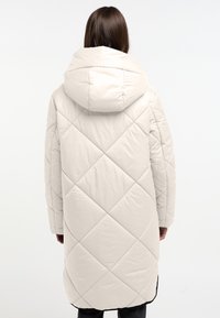 Lange, beige pufferjas met een diamanten quiltpatroon, voorzien van een grote capuchon en lange mouwen. De onderrand heeft een zwarte accentrand.